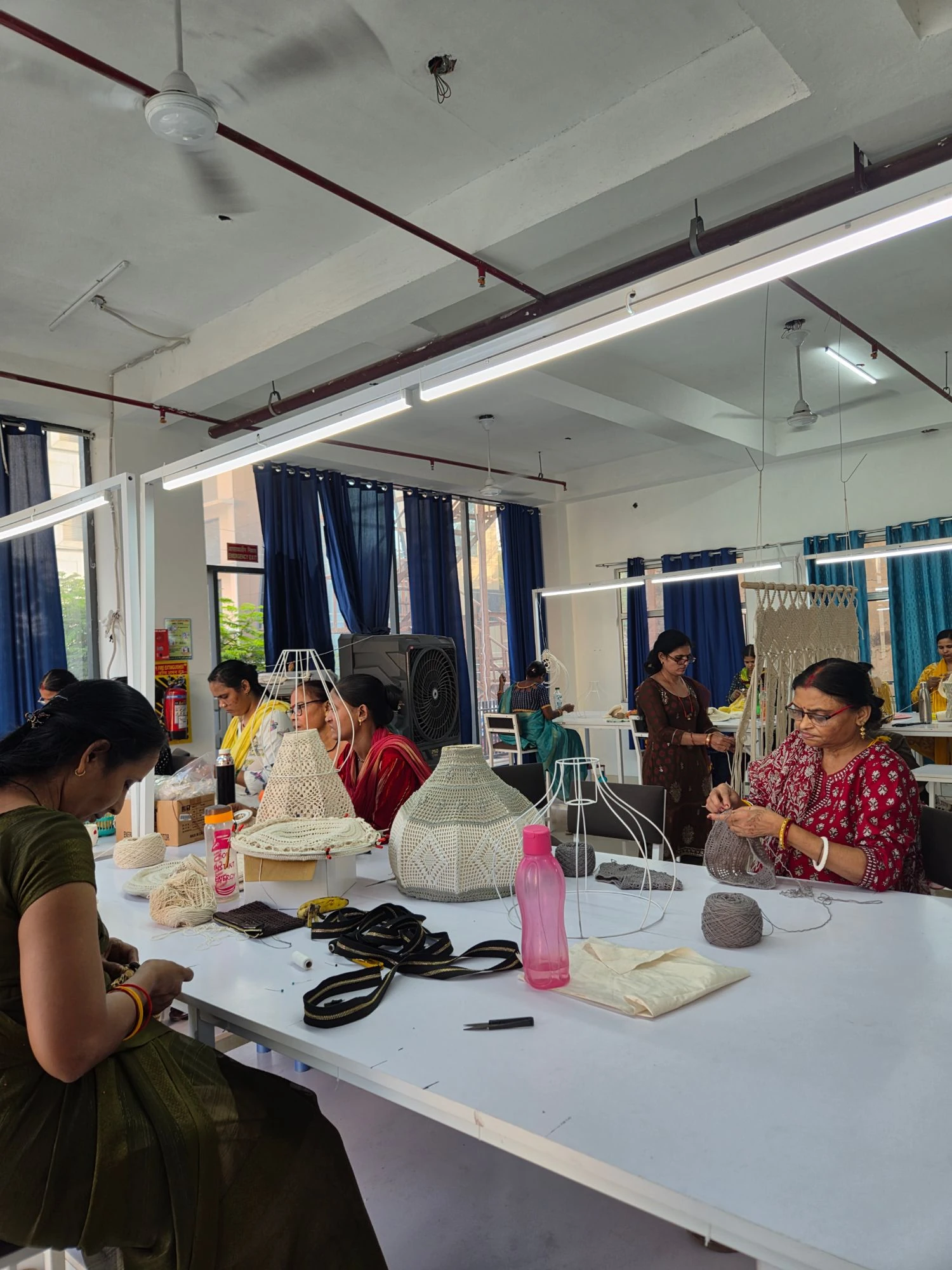Makers crocheting lampshades and bags at long white worktables on our production floor, with lamp frames and yarn on the tables.