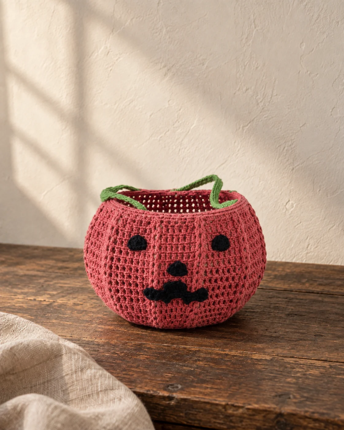 Pink crochet pumpkin-face basket with black facial features and a green handle.