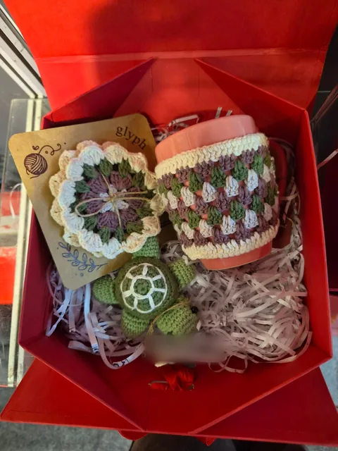A red gift box holding a crochet flower coaster, a mug in a crochet cosy and a crochet turtle, with a Glyph by Vera Verve card, at our stall, Gifts World Expo 2026.