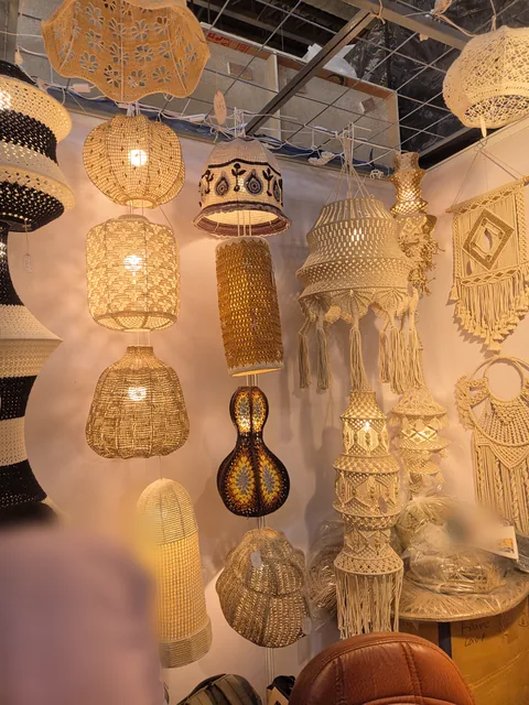 A corner of our stall at IHGF Delhi Fair Autumn 2026 hung with crochet and macramé lamps: round and tall cream shades, a black and cream patterned shade, a brown gourd-shaped lamp and tasselled macramé pendants beside a macramé diamond wall hanging.