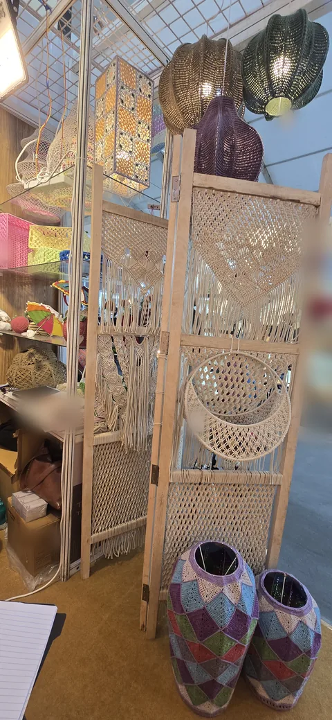 A wooden folding screen with macramé panels and a hanging macramé shelf, two crochet vases in purple, green and pink below it and crochet pendant lamps above, at our stall, IHGF Delhi Fair Spring 2025.