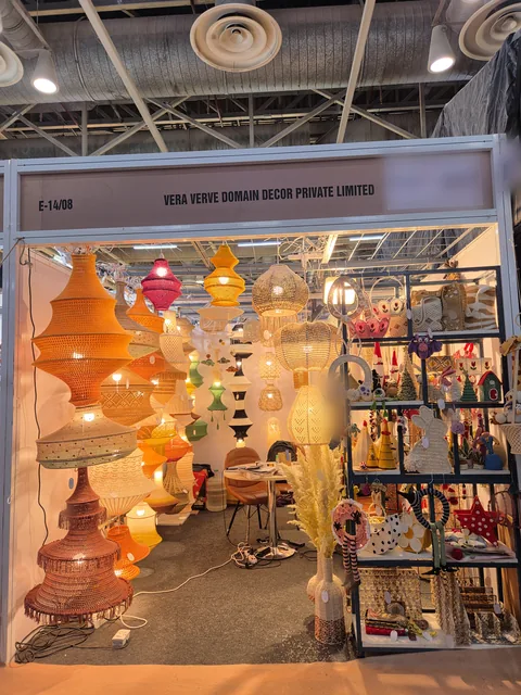 The front of our stall E-14/08 at IHGF Delhi Fair Autumn 2026, with tall orange and rust crochet pendant lamps on the left and shelves of crochet toys and baskets on the right.