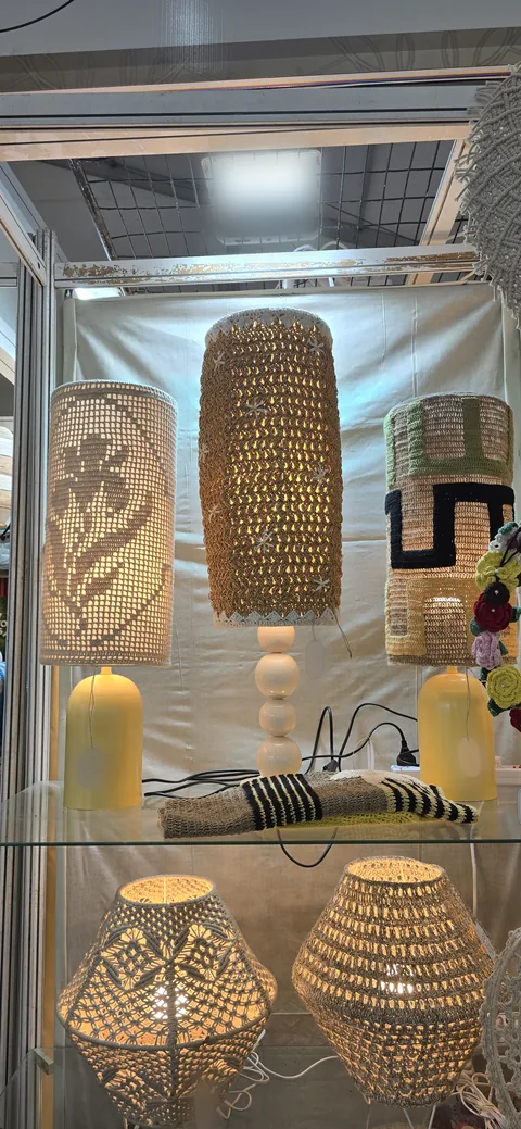 Three crochet table lamps with tall cylinder shades on yellow and white bases, above two lit round crochet lamps on a glass shelf at our stall, IHGF Delhi Fair Autumn 2025.
