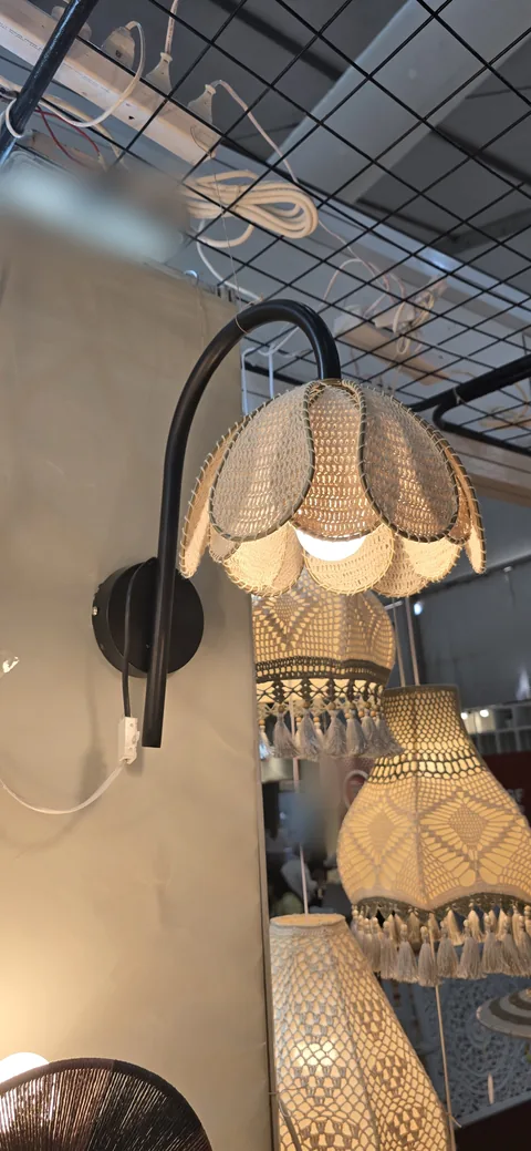 A wall lamp with a scalloped crochet shade on a curved black arm, with tasselled crochet pendant lamps behind it at our stall, IHGF Delhi Fair Autumn 2025.