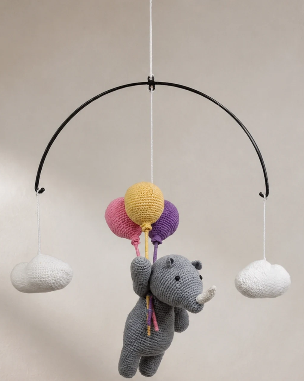 Crocheted grey rhino holding pink, yellow and purple balloons, hanging between two white clouds.